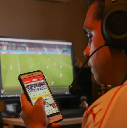 Un homme portant un casque sur la tête, qui est sur le point de parier avec son téléphone sur un match de foot qu'il est entrain de regarder sur son ordinateur